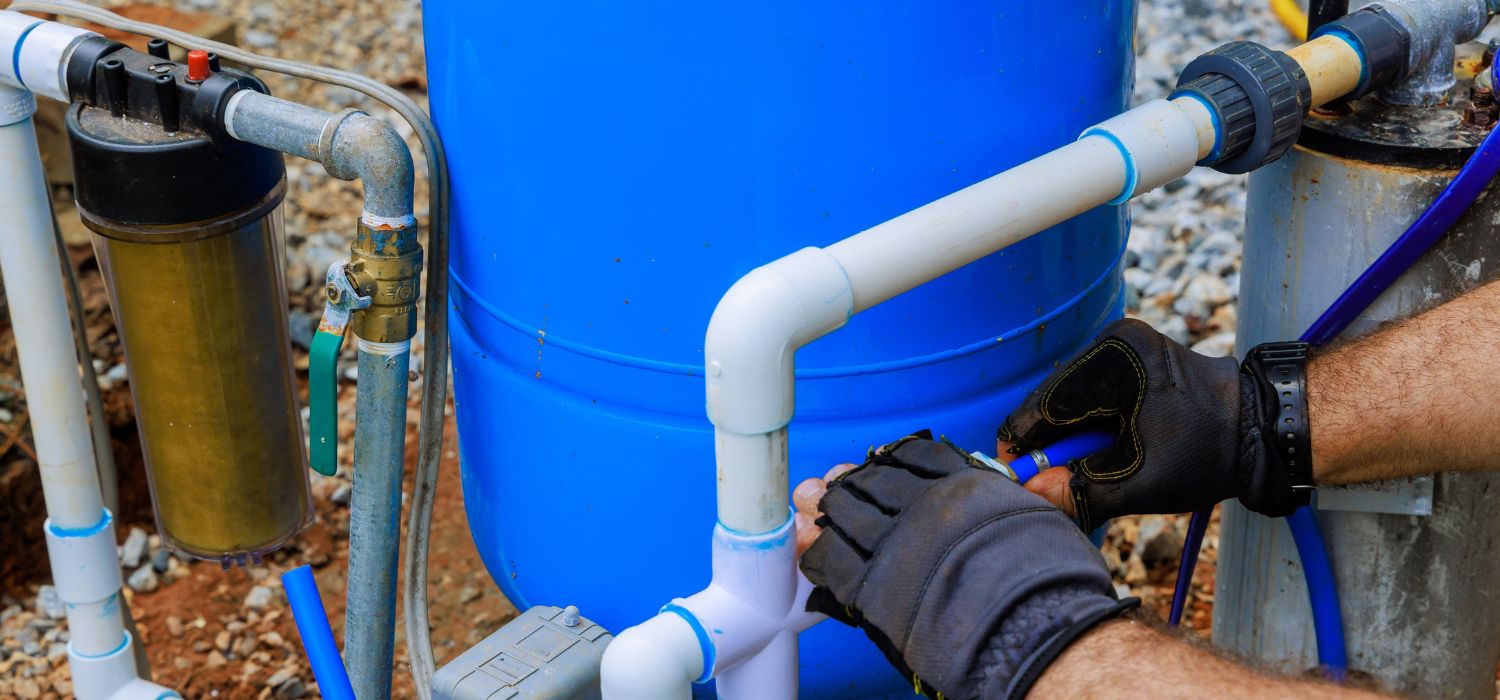 Technician servicing a residential well pump system with pressure tank, filtration unit, and PVC plumbing connections during maintenance.