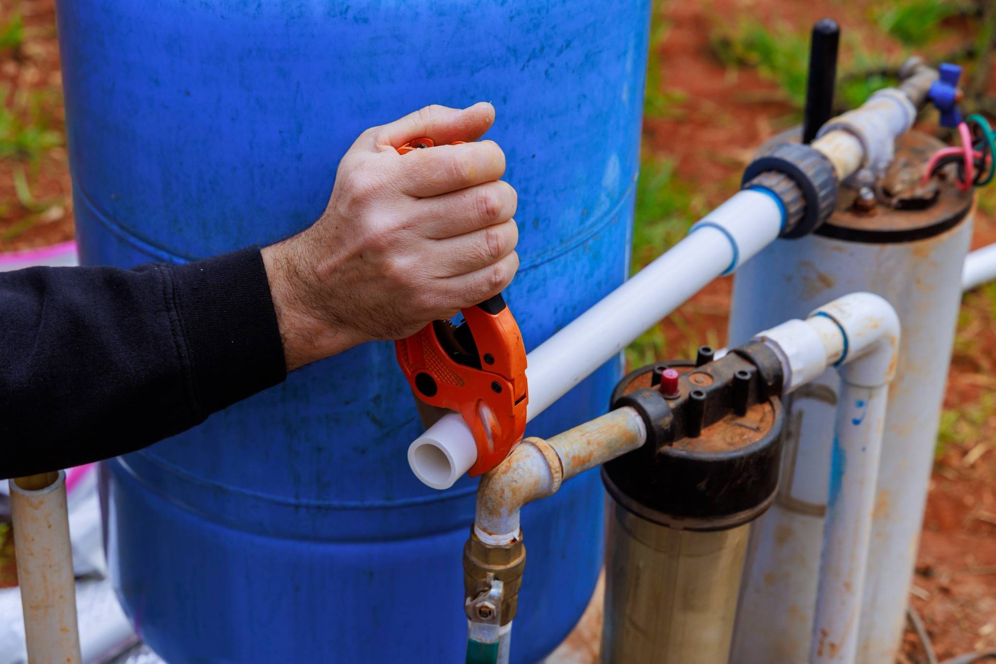Person using a pipe cutter to cut PVC pipe connected to a well pump system with blue water tank and filtration components.