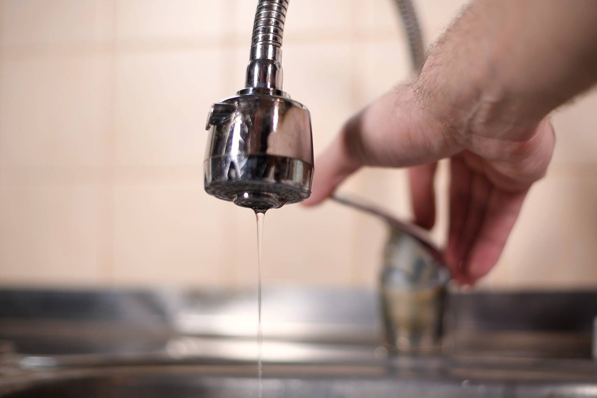 A hand turns a kitchen faucet as a thin stream of water drips, highlighting concerns about water quality and well water safety.